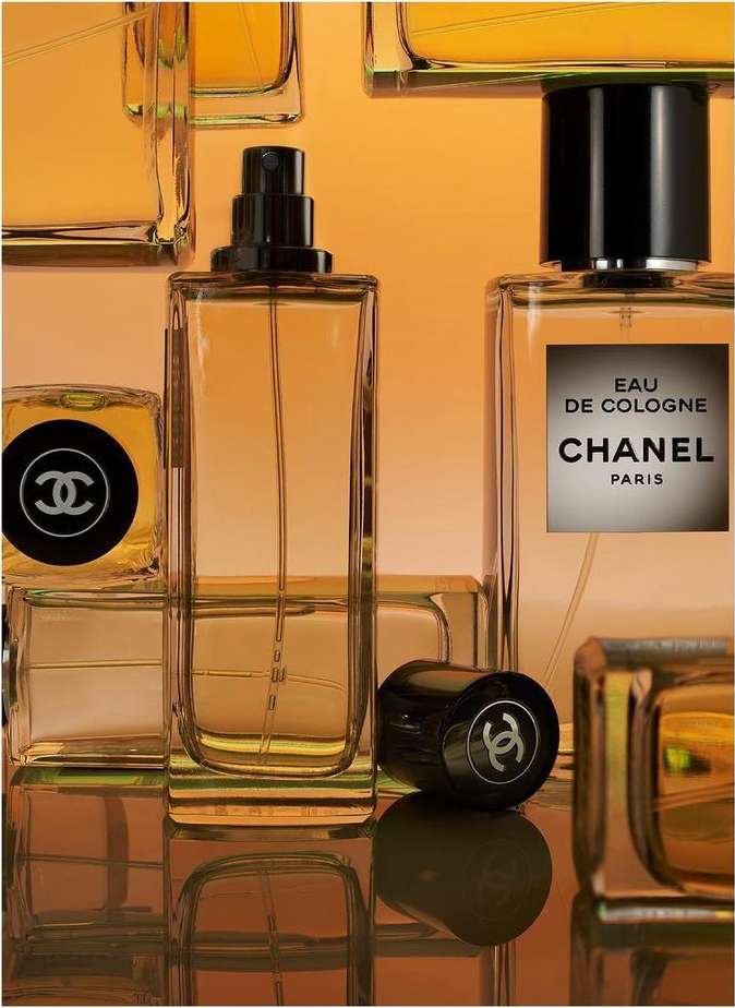 Several clear Chanel cologne bottles sit on a reflective surface against an orange backdrop, showcasing minimalist design and luxury branding.