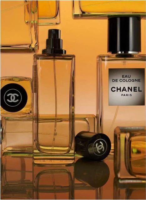 Several clear Chanel cologne bottles sit on a reflective surface against an orange backdrop, showcasing minimalist design and luxury branding.