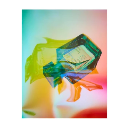 An artistic, abstract product shot features an emerald glass perfume bottle with soft gradients of green, yellow, orange, and pink.