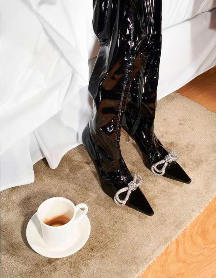 Over-the-knee patent leather boots with decorative bows, set on a beige rug near a coffee cup and a bed with white sheets.