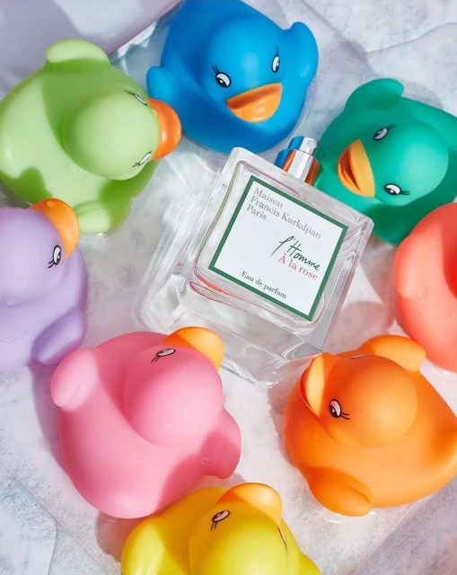 Square bottle of Maison Francis Kurkdjian Paris A la rose perfume surrounded by colorful rubber ducks on white crinkled material.