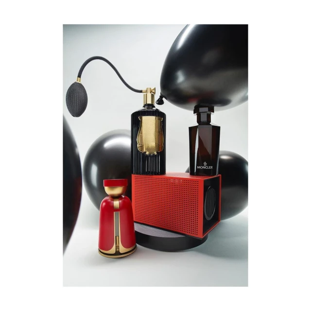 Still life studio shot features two perfume bottles, a speaker, and decorative figure against a gradient white backdrop, accentuated by black balloons.