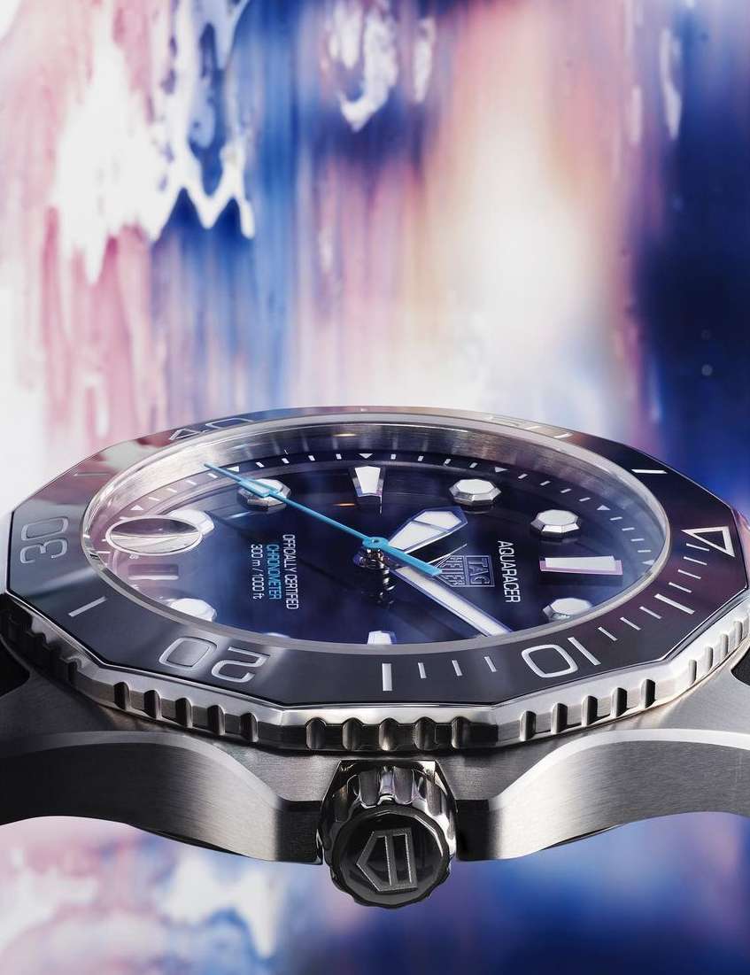 Close-up of a stainless steel Tag Heuer Aquaracer watch with a blue face against a blurred background of blue and pink hues.