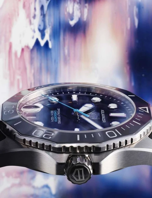 Close-up of a stainless steel Tag Heuer Aquaracer watch with a blue face against a blurred background of blue and pink hues.