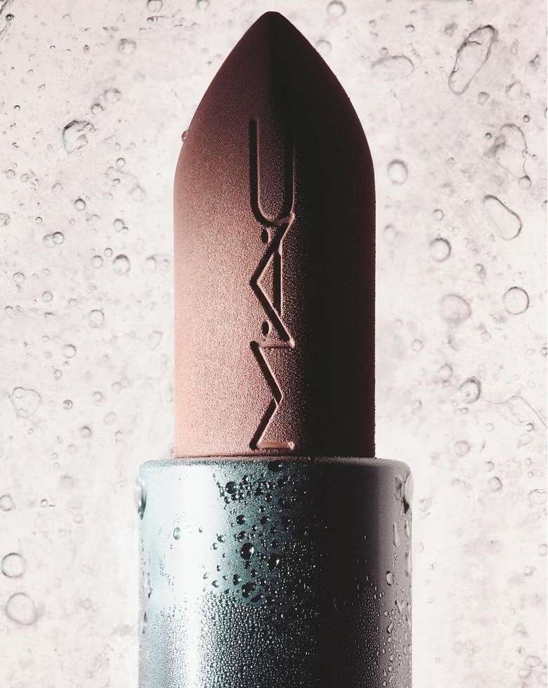 Close-up product shot of a brown MAC lipstick with water droplets. The lipstick has the MAC logo embossed and is set against a blurred backdrop.