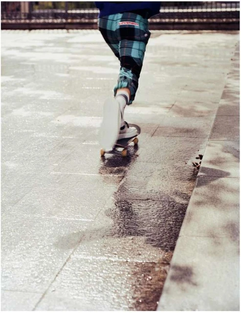 Person skateboarding in a city environment on a wet surface, wearing plaid pants and white sneakers, with a stone sidewalk.