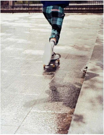 Person skateboarding in a city environment on a wet surface, wearing plaid pants and white sneakers, with a stone sidewalk.