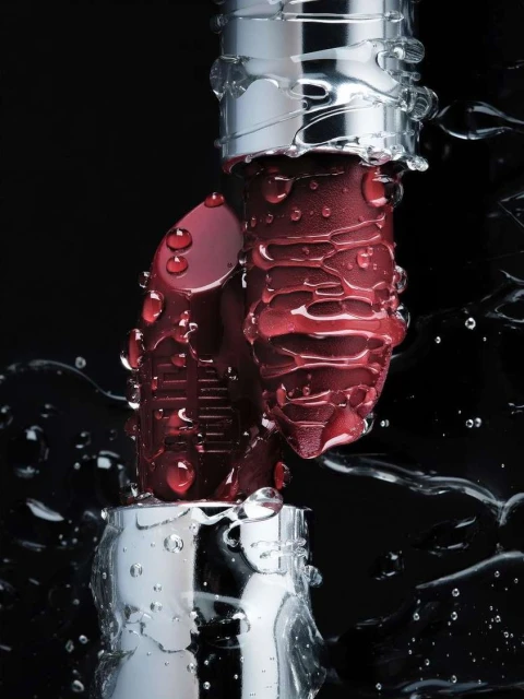 Close-up product shot of two burgundy lipsticks with silver casings covered in water droplets against a dark black background.
