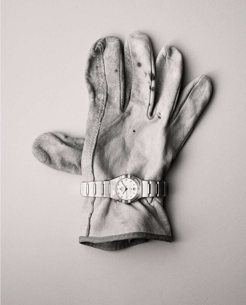 A glove with a watch on top of a plain background. The leather work glove is worn, and the watch has a silver metal band.