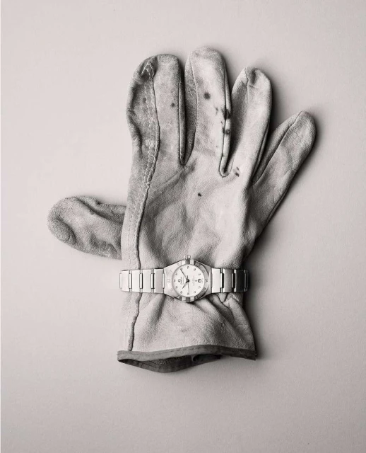 A glove with a watch on top of a plain background. The leather work glove is worn, and the watch has a silver metal band.