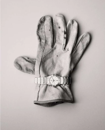 A glove with a watch on top of a plain background. The leather work glove is worn, and the watch has a silver metal band.
