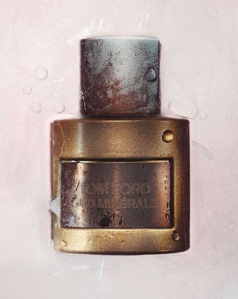 Close-up studio shot of Tom Ford Oud Minerale perfume bottle with wet surface and pink background, showcasing textures and details.