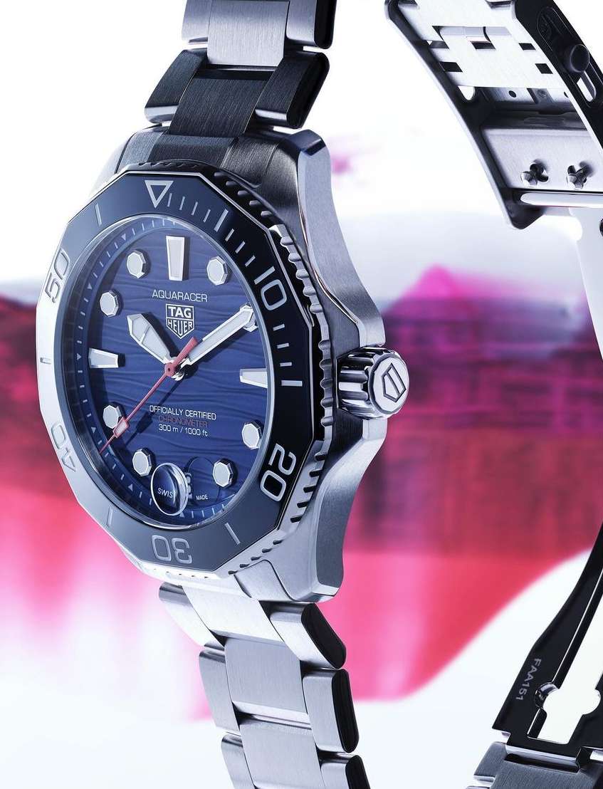 Silver Tag Heuer Aquaracer wristwatch with a blue dial on a pink and white blurred background, product shot.