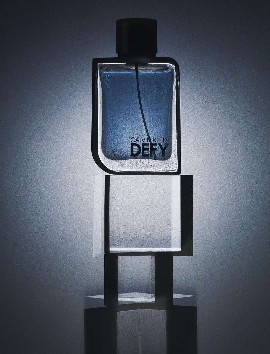 Calvin Klein Defy perfume bottle displayed atop a clear, geometric stand with a blue and gray color scheme and minimalist style.