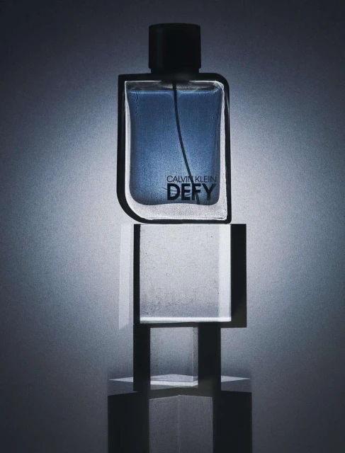 Calvin Klein Defy perfume bottle displayed atop a clear, geometric stand with a blue and gray color scheme and minimalist style.