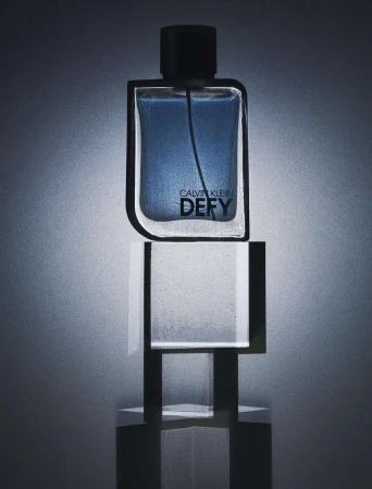 Calvin Klein Defy perfume bottle displayed atop a clear, geometric stand with a blue and gray color scheme and minimalist style.