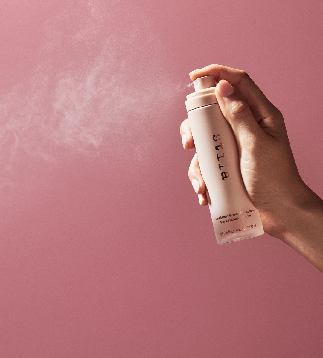Close-up studio shot of a light-skinned hand holding and spraying a tan setting spray bottle against a pink background.