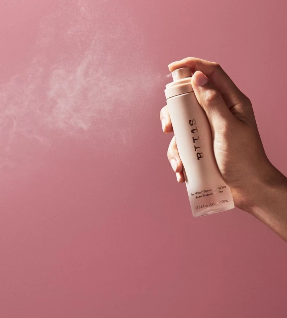 Close-up studio shot of a light-skinned hand holding and spraying a tan setting spray bottle against a pink background.