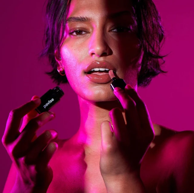 Close-up of an adult woman applying Pavise lip gloss, bathed in pink light with a soft focus and shallow depth of field.
