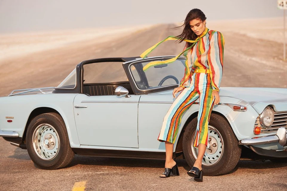 A brunette woman in a colorful striped outfit leans against a light blue vintage convertible with a road and tan hills in the background.