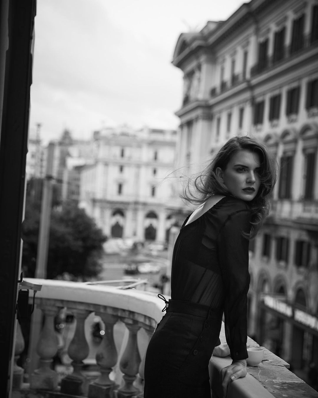 Black and white medium shot of a woman posing on a balcony with an urban background featuring buildings and city street.