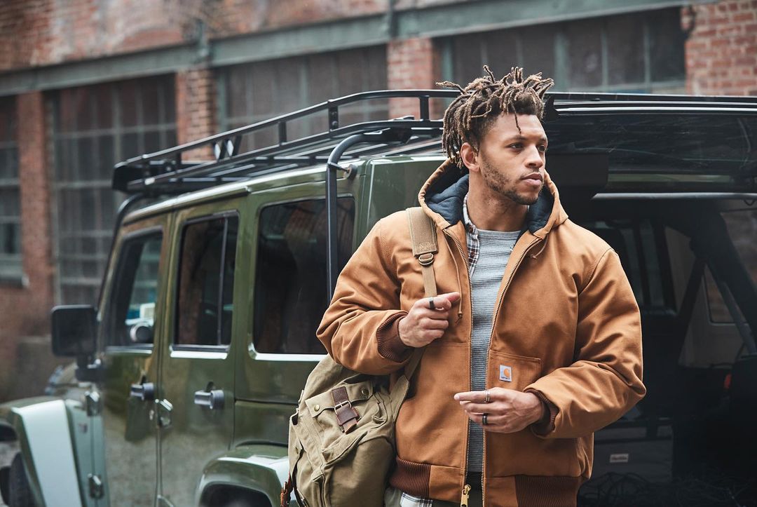 Adult male with dreadlocks in a Carhartt jacket stands near an olive green SUV on an urban street with brick buildings in the background.
