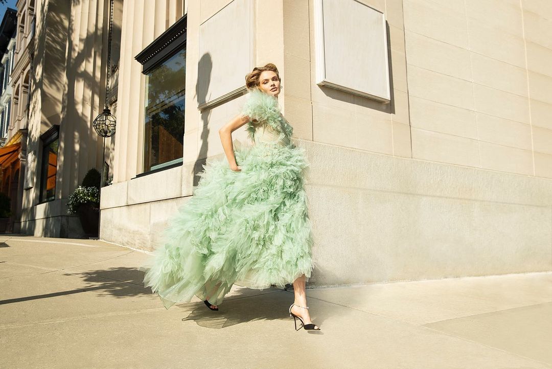 Fashionable adult woman in a light green ruffled gown, posing on an urban street with neutral colored buildings on a sunny day.