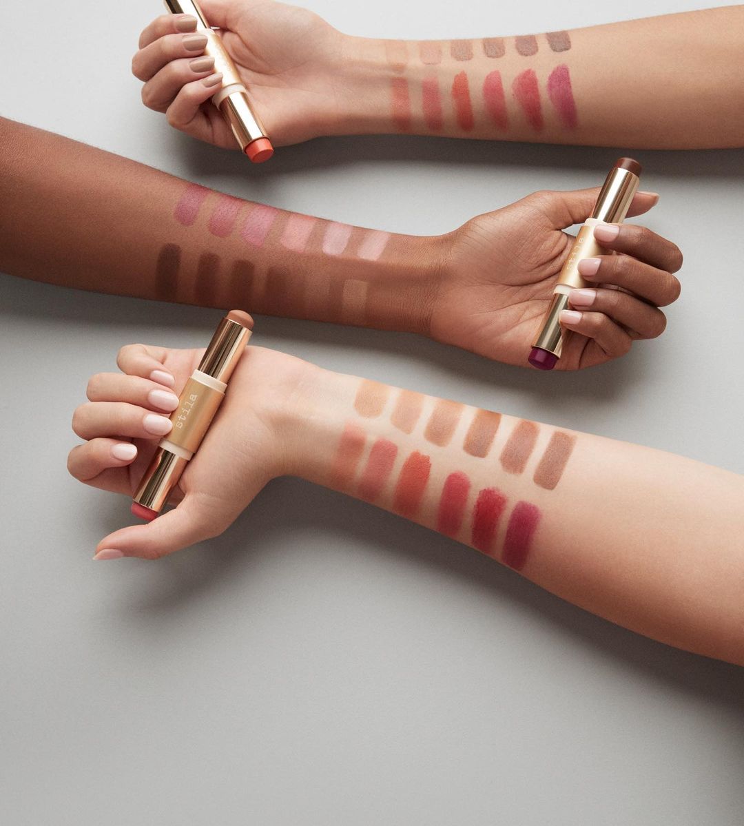 Three arms display makeup swatches and a dual-ended contour stick on a gray background, showcasing beauty and cosmetic artistry.