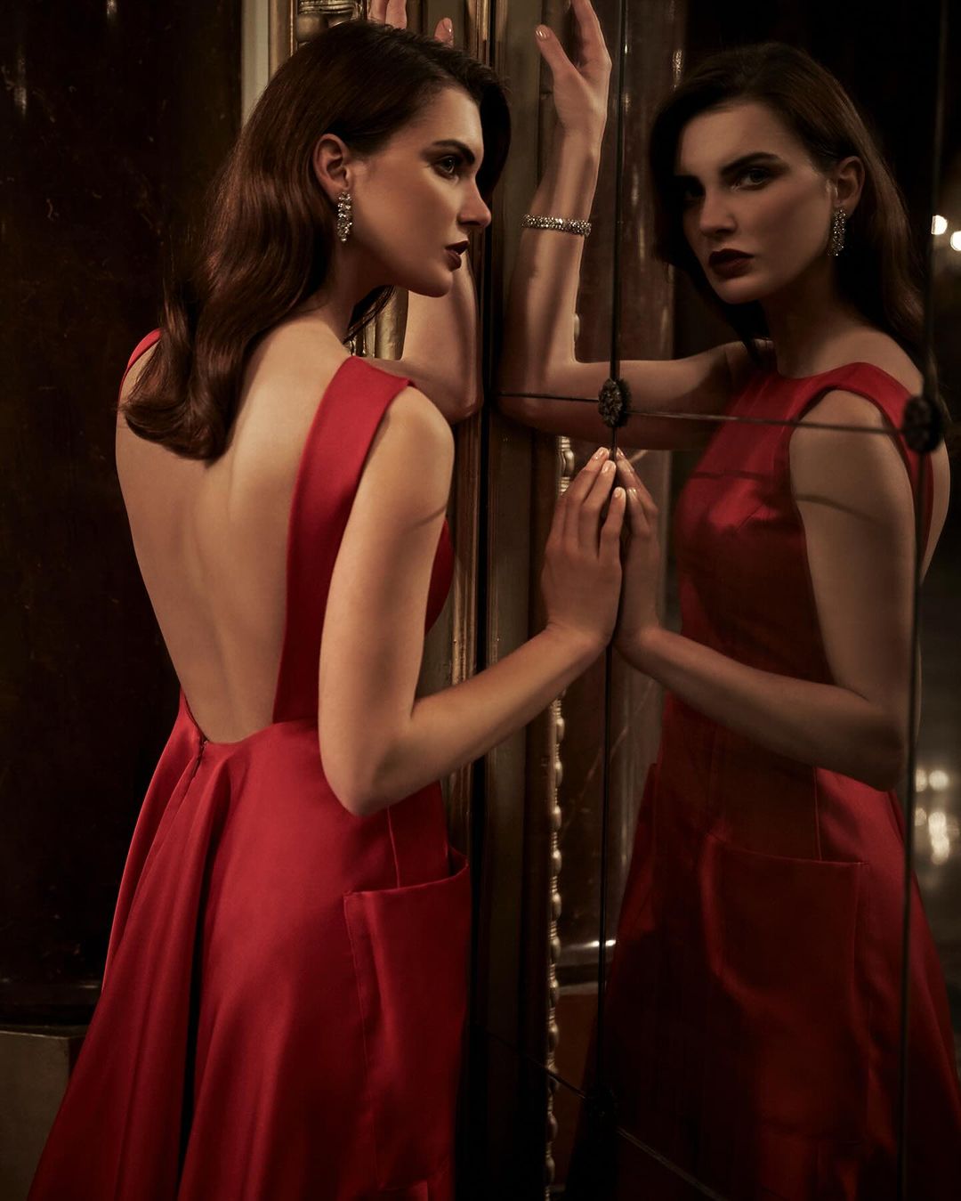 Elegant woman in a red gown poses with a mirror, showing her reflection. Dark hair and sophisticated style.