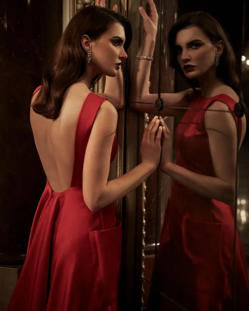 Elegant woman in a red gown poses with a mirror, showing her reflection. Dark hair and sophisticated style.