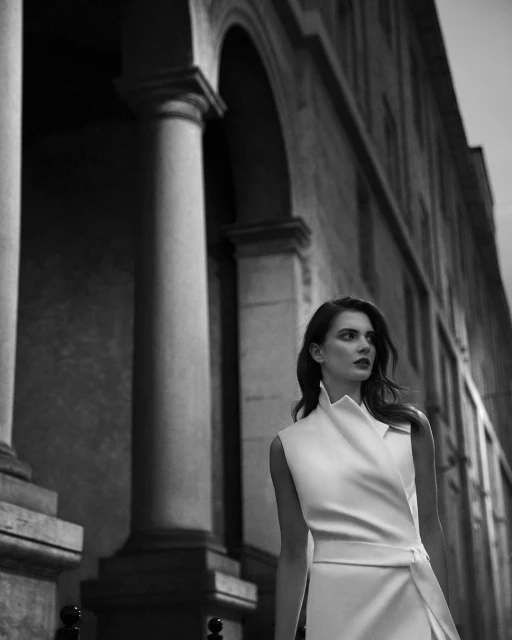 Monochrome portrait of a stylish woman in a white sleeveless dress, standing against a classical architectural backdrop with stone columns.