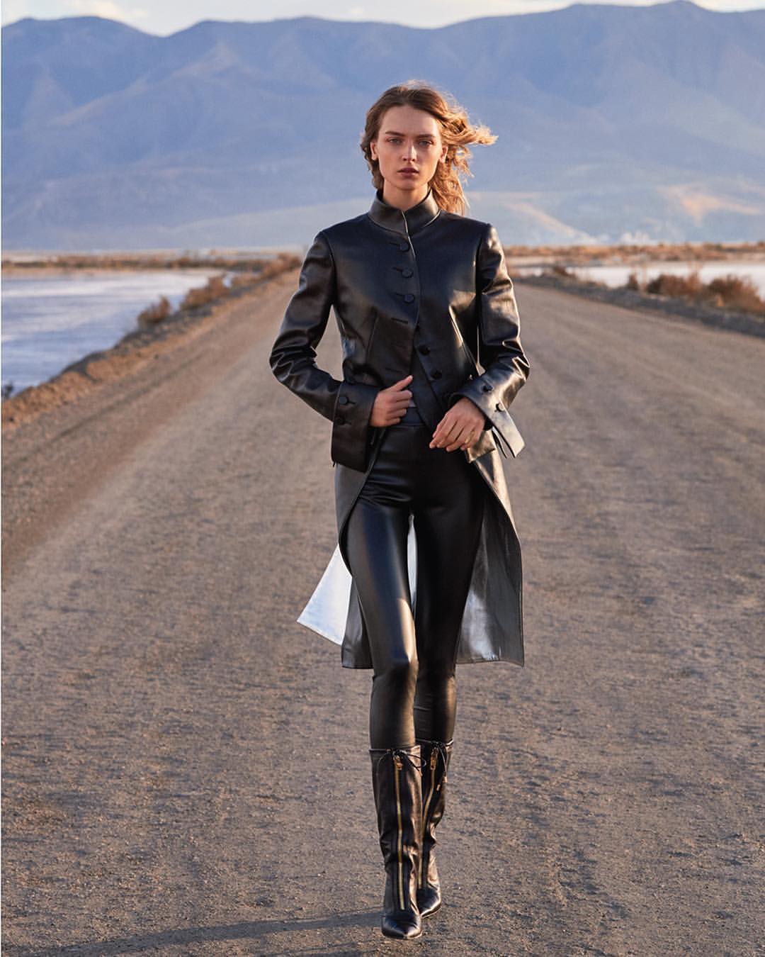 Woman in black leather outfit confidently walks down a desert road toward the camera, mountains in background, outdoor fashion shot.