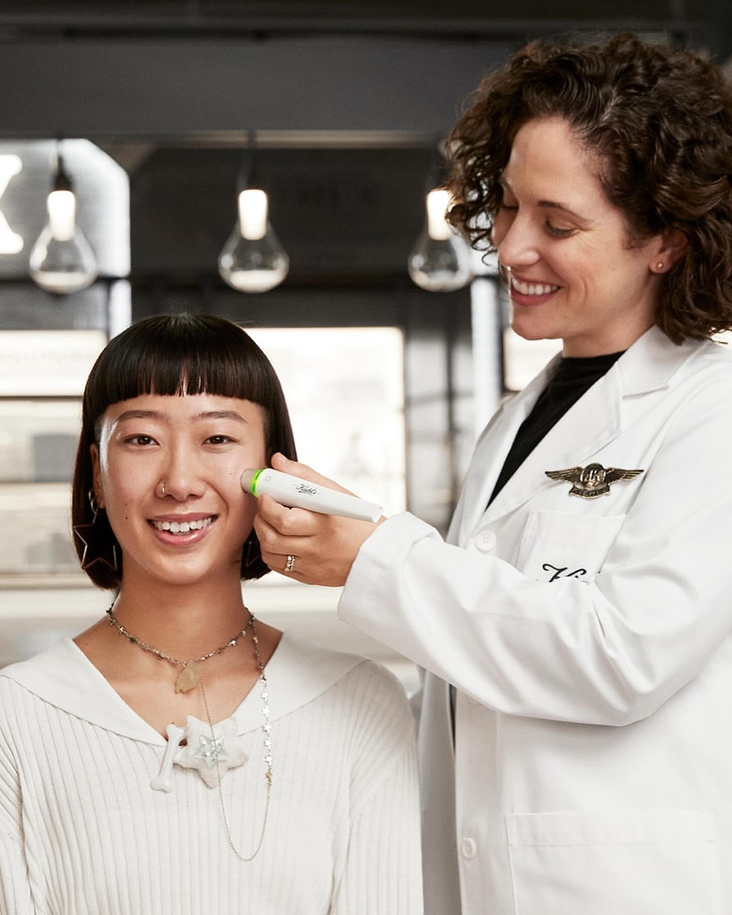 Smiling Asian woman receives a facial treatment from a doctor in a white coat indoors, focusing on skincare and beauty.