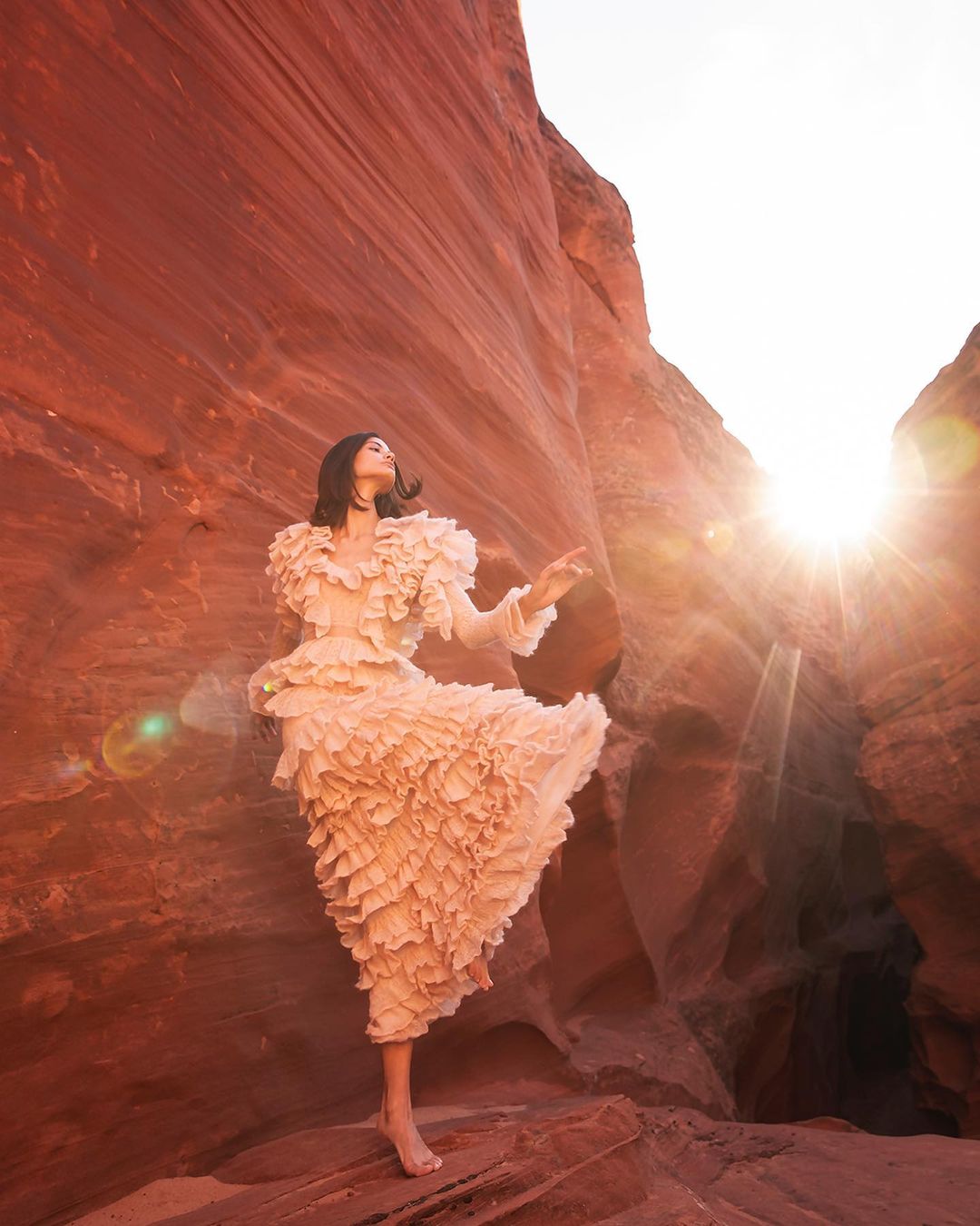 Woman in ruffled dress poses barefoot among red rock formations in a sunlit canyon, creating a striking fashion editorial photograph.
