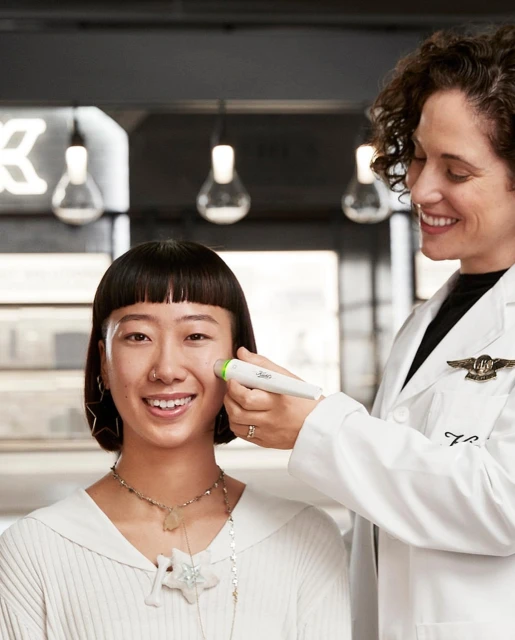 Asian woman smiles during facial treatment. A beautician in a white coat uses a wand on her cheek in an indoor environment.