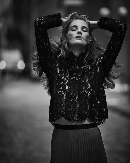 Black and white portrait of a blonde female model posing with closed eyes in lace top and pleated skirt on a city street at night.