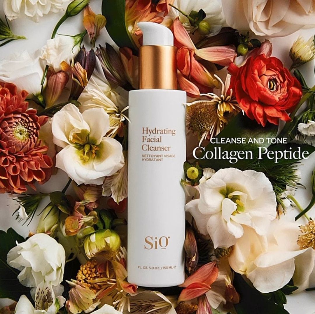A white bottle of Si0 Hydrating Facial Cleanser with a gold pump surrounded by a variety of colorful flowers in a close-up shot.