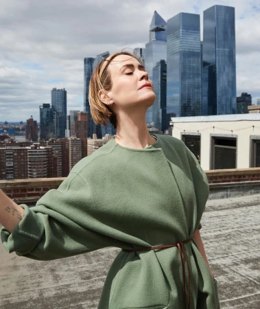 Sarah Paulson wearing an olive green coat outdoors on a New York rooftop against a city skyline