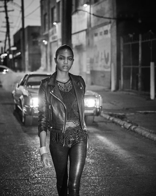 Monochrome fashion shot of a woman in a leather jacket walking on a street with an automobile in the background at night.