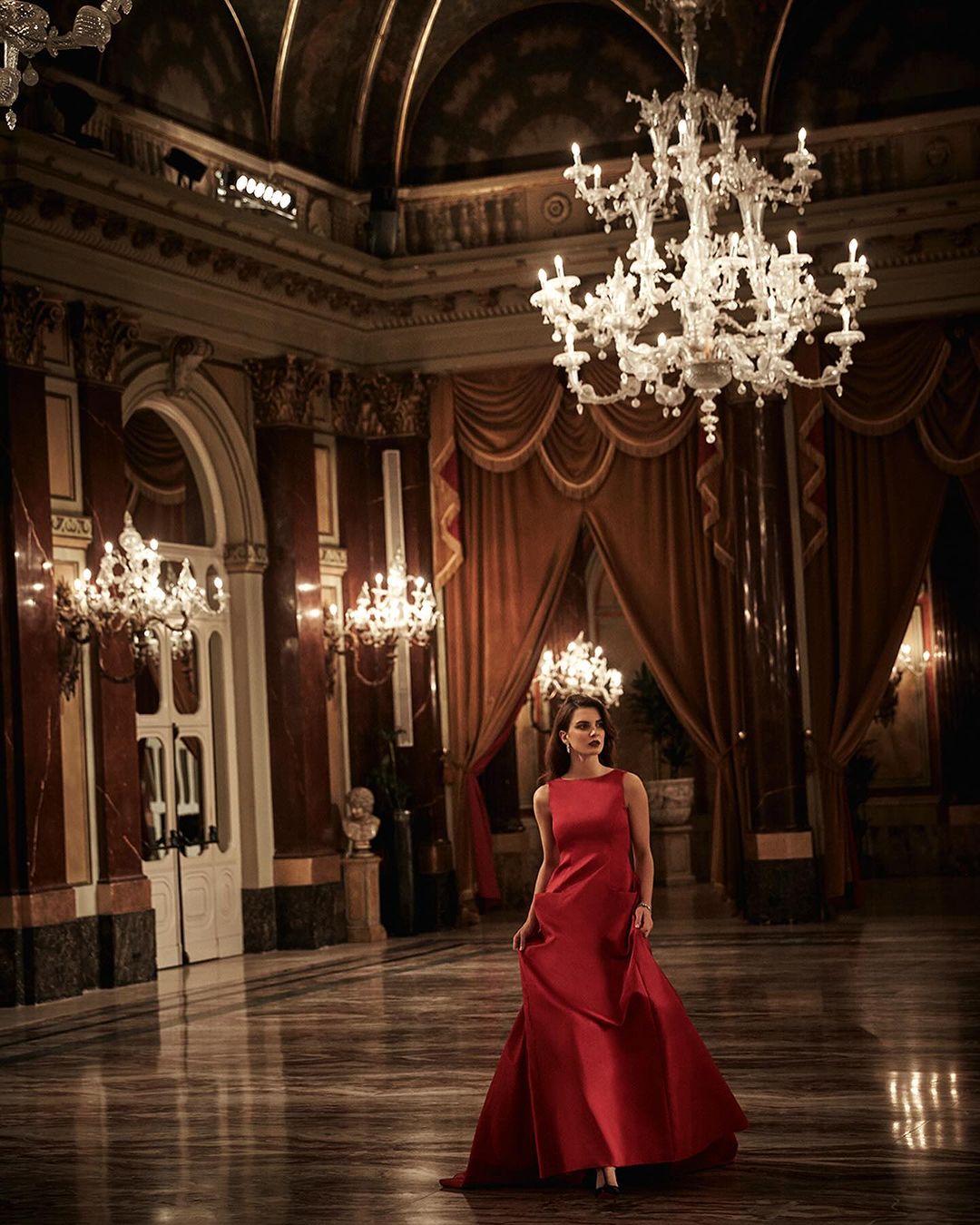 Elegant woman in a red gown stands in a grand ballroom beneath a chandelier, surrounded by ornate architecture and marble flooring.