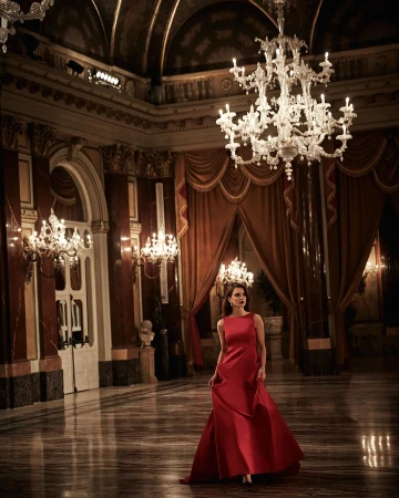 Elegant woman in a red gown stands in a grand ballroom beneath a chandelier, surrounded by ornate architecture and marble flooring.