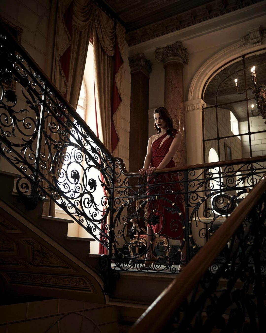 Woman in red dress on ornate staircase. Dark, moody, low-angle shot inside mansion with marble columns and decorative ironwork.