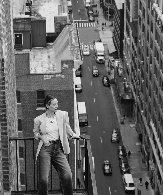 Black and white image of a woman in a blazer and jeans standing on a rooftop overlooking a city street with buildings and traffic.