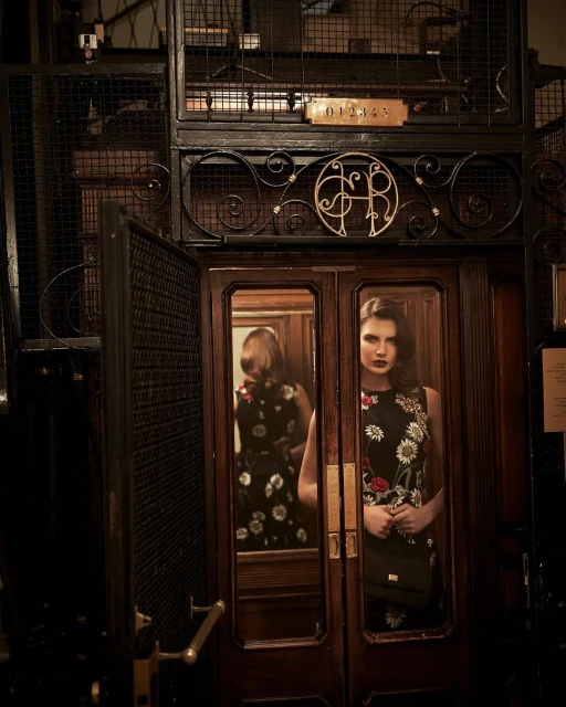 Woman in floral dress in vintage elevator with ornate metalwork and dark wood details, showcasing fashion photography in indoor setting.