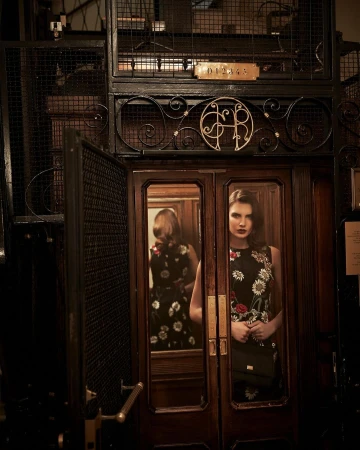 Woman in floral dress in vintage elevator with ornate metalwork and dark wood details, showcasing fashion photography in indoor setting.