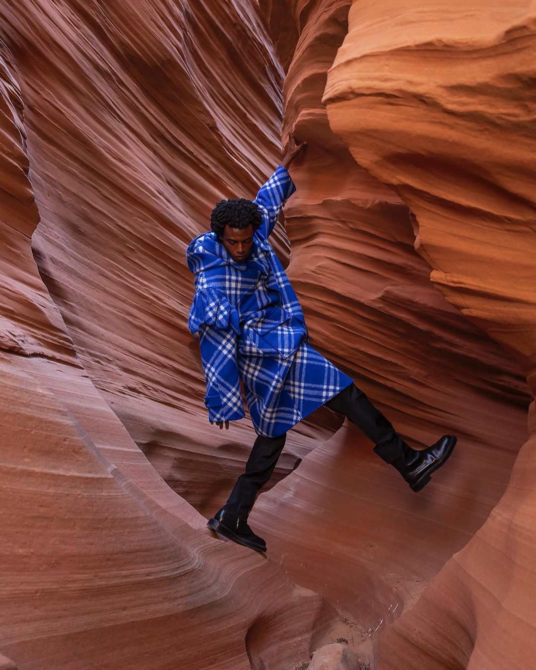 A male model in a blue plaid coat poses dynamically within a slot canyon, wearing black pants and boots, surrounded by textured sandstone.