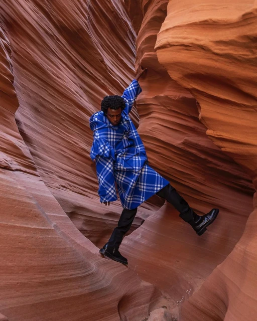 A male model in a blue plaid coat poses dynamically within a slot canyon, wearing black pants and boots, surrounded by textured sandstone.