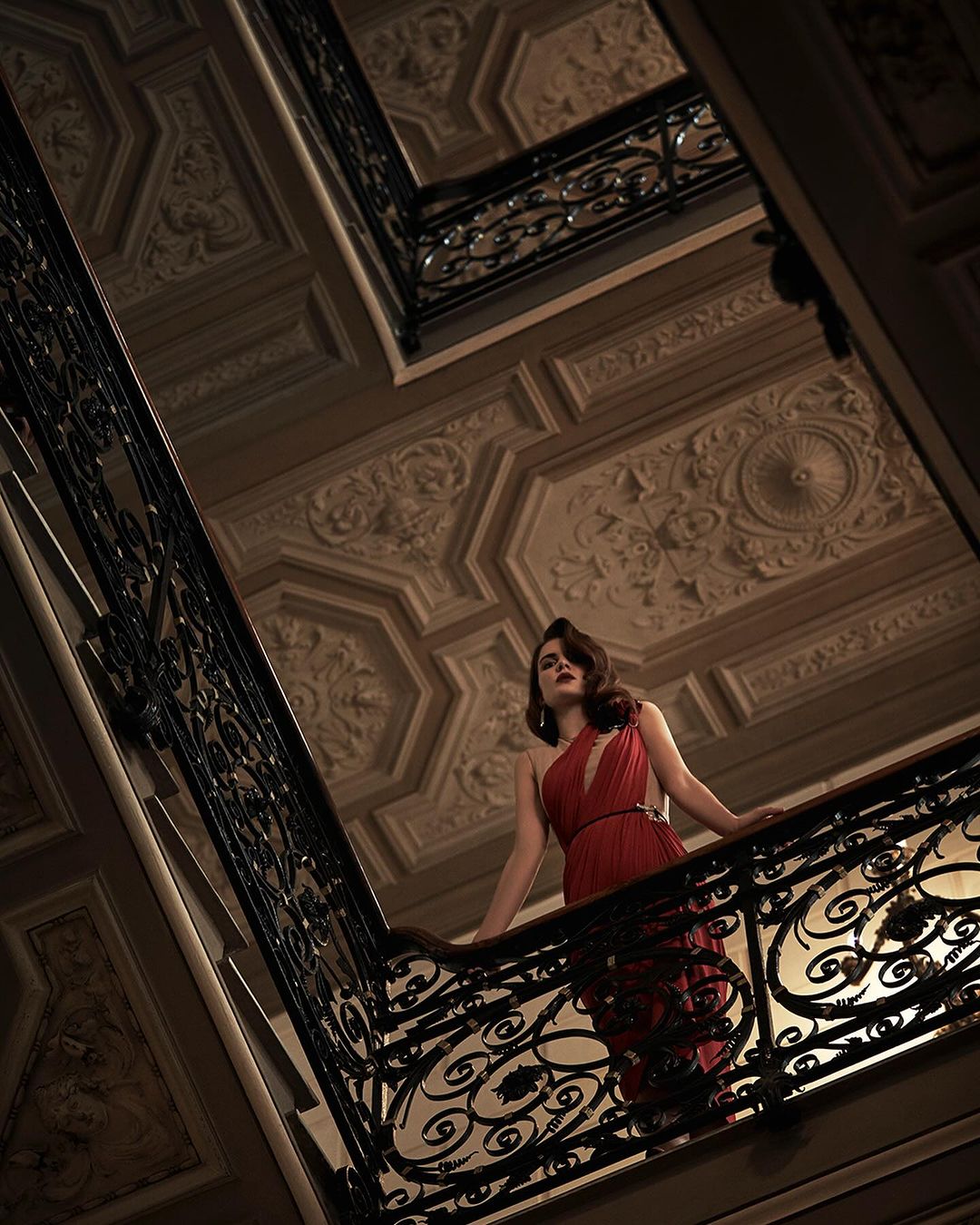 Elegant woman in red dress poses on ornate wrought-iron balcony. Interior setting with decorative plaster ceiling, low-angle shot.