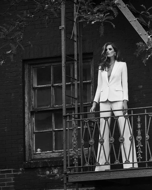 Monochrome portrait of a stylish adult woman posing in a white pantsuit on an outdoor balcony with a fire escape and brick wall.