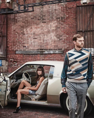 Man standing foreground in patterned sweater and woman seated inside vintage car against brick wall by Tom Corbett